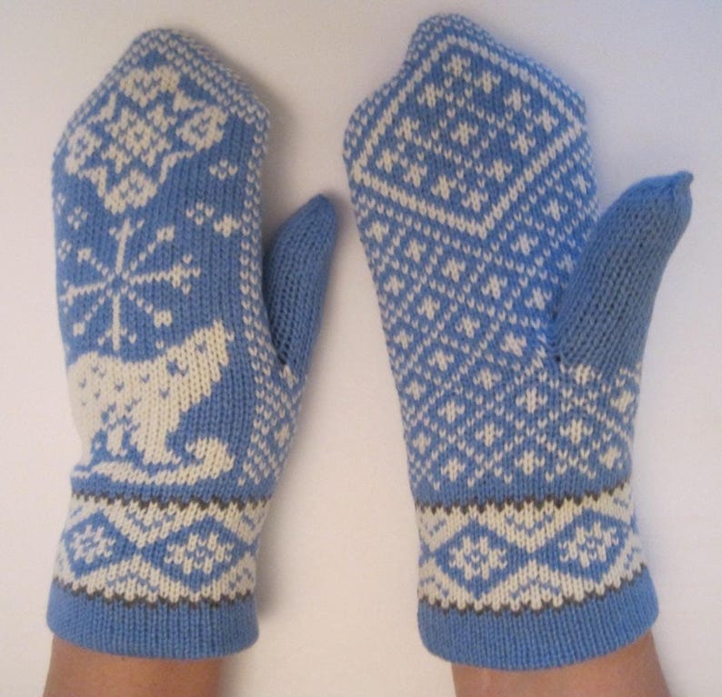 May include: A pair of blue knitted mittens with white and brown geometric and animal patterns. The mittens feature a snowflake and polar bear design on one and a diamond pattern on the other. The cuffs and thumbs are solid blue.