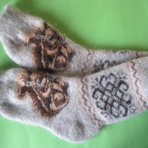 May include: A pair of gray wool socks with brown and black patterned designs. The socks feature a repeating pattern of a brown animal on the front and a black and brown geometric design on the back. The socks are on a green background.