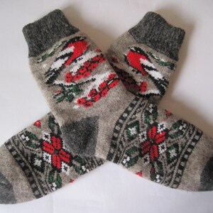 May include: A pair of gray wool socks with a bird and floral design. The socks feature a red bird, red berries, and green leaves on the upper portion. The lower portion has a red and white floral pattern with green accents.