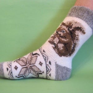 Puede incluir: Calcetines de lana blanca con un diseño de ardilla marrón y un puño gris. Los calcetines tienen un patrón de copo de nieve en el pie.