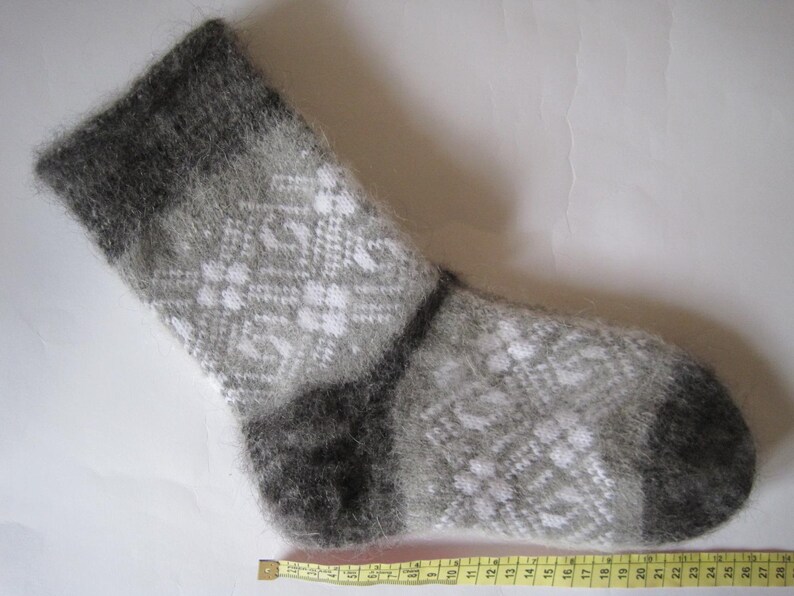 K&ouml;nnte beinhalten: Ein flauschiger, wadenhoher Socken mit einem grau-wei&szlig;en geometrischen Rautenmuster. Der Socken hat ein dunkelgraues B&uuml;ndchen sowie eine dunkelgraue Ferse und Spitze. Ein gelbes Ma&szlig;band liegt darunter und zeigt eine L&auml;nge von etwa 28 cm.