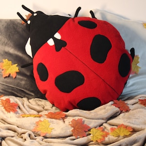 Op de afbeelding: Een pluche kussen in de vorm van een lieveheersbeestje met een felrood lichaam, zwarte ronde stippen en een zwart kopgedeelte. Het kussen heeft kleine zwarte voelsprieten en pootjes aan de zijkanten, gemaakt van een zachte, textuurrijke stof.