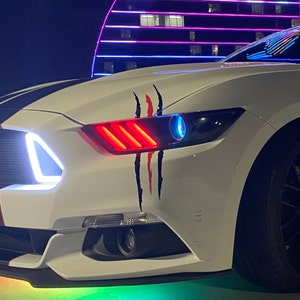 Puede incluir: Un Ford Mustang blanco con rayas de carreras negras y faros LED rojos y azules. El coche tiene una calcomanía de garra negra y roja en el guardabarros delantero.