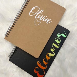 May include: Two spiral bound notebooks. One is brown with the name "Olivia" written in white with a white heart. The other is black with the word "cleaners" written in rainbow colors.