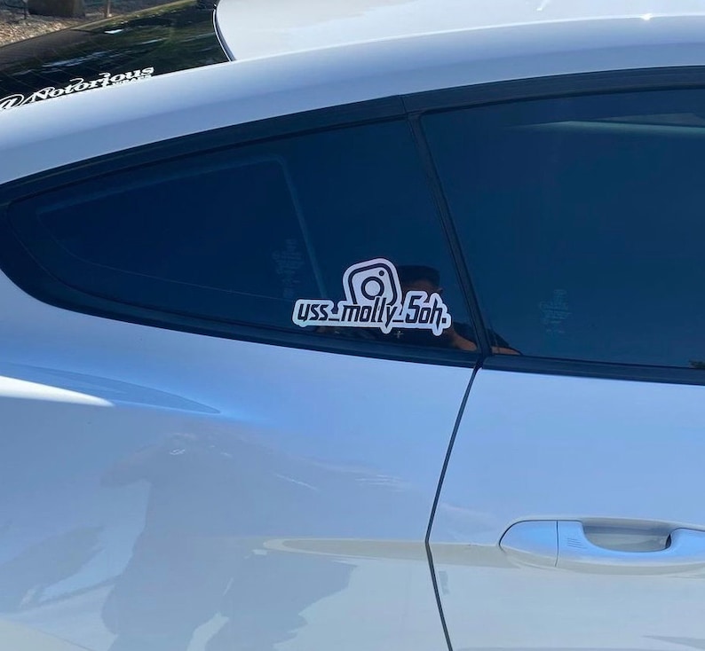 May include: A white vinyl decal applied to the rear side window of a silver car. The decal features a stylized Instagram camera icon positioned above the text "yss_molly5oh" in a bold, sans-serif font with a thick white outline and black fill.