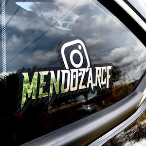 May include: A custom decal applied to a vehicle window featuring a carbon fiber texture background. The design includes a white Instagram logo icon positioned above the text MENDOZA.RCF, which is rendered in a bold, stylized green camouflage font.