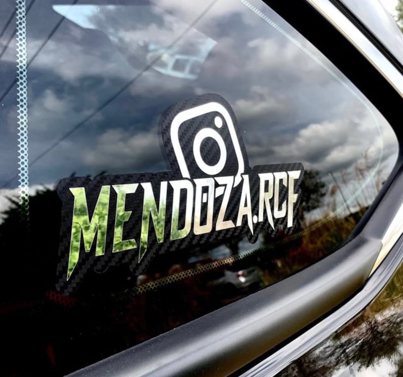 May include: A custom decal applied to a vehicle window featuring a carbon fiber texture background. The design includes a white Instagram logo icon positioned above the text MENDOZA.RCF, which is rendered in a bold, stylized green camouflage font.