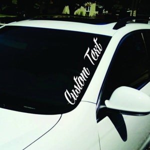 May include: White car with a windshield banner. The banner features the words "Custom Text" in a cursive font. The car is parked outside on a sunny day.