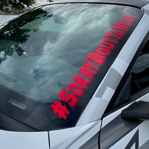 Puede incluir: Una calcoman&iacute;a de vinilo roja aplicada en la parte superior del parabrisas de un autom&oacute;vil. La calcoman&iacute;a presenta el texto #SORRYBOUTYANECK en una fuente negrita sin remates, colocada diagonalmente sobre el cristal del veh&iacute;culo blanco.