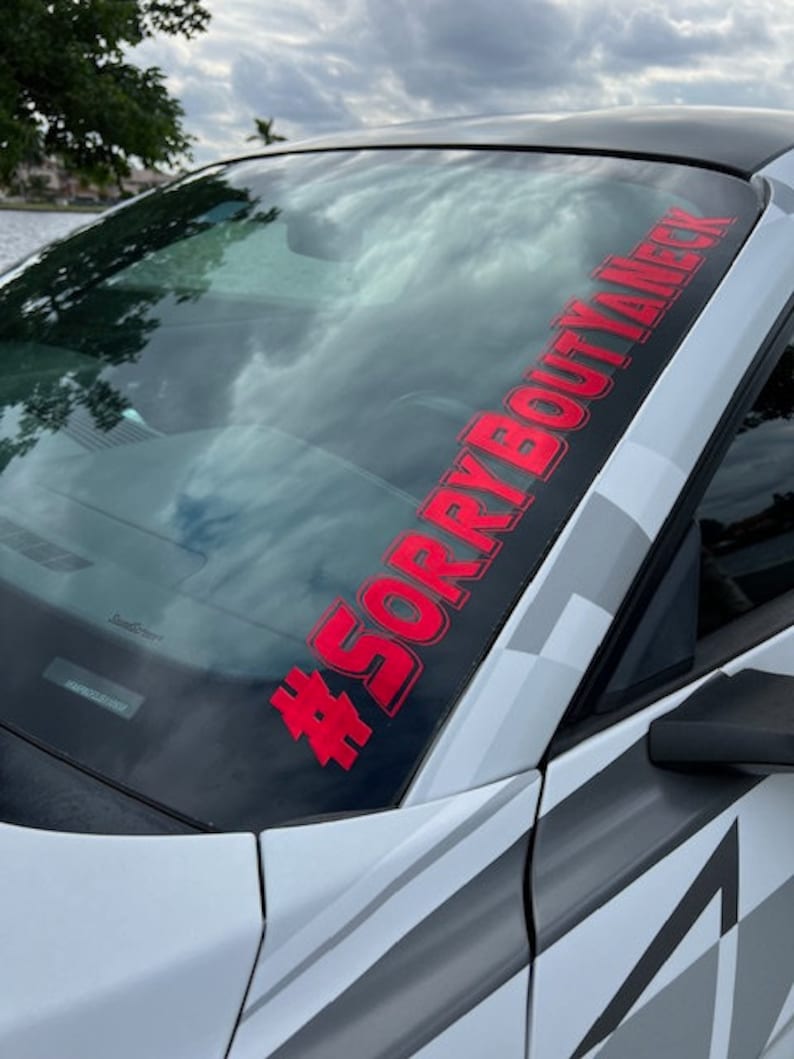 Puede incluir: Una calcoman&iacute;a de vinilo roja aplicada en la parte superior del parabrisas de un autom&oacute;vil. La calcoman&iacute;a presenta el texto #SORRYBOUTYANECK en una fuente negrita sin remates, colocada diagonalmente sobre el cristal del veh&iacute;culo blanco.