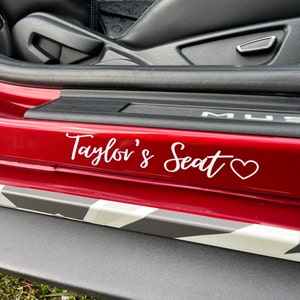 Op de afbeelding: Een rode autodeur met een witte vinyl sticker waarop "Taylor's Seat" staat met een hartje.