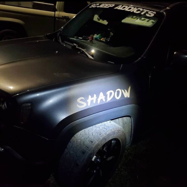 Puede incluir: Un veh&iacute;culo gris oscuro con acabado mate que presenta el texto blanco SHADOW en el guardabarros delantero y SC JEEP ADDICTS en el parabrisas. El veh&iacute;culo tiene ruedas negras y neum&aacute;ticos texturizados, captado con poca luz y un reflejo.
