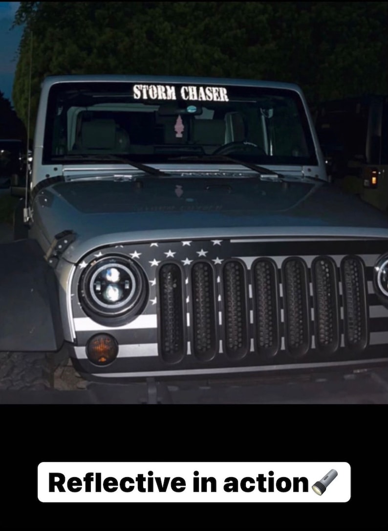 Puede incluir: Un veh&iacute;culo Jeep plateado con un gr&aacute;fico de la bandera estadounidense en blanco y negro sobre la parrilla delantera. El parabrisas muestra una calcoman&iacute;a blanca con el texto STORM CHASER. Los faros circulares est&aacute;n encendidos y cuelga un ambientador.