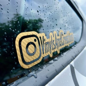 May include: A gold and black Instagram logo sticker with the text "Vinyl's And Creations" on a car window with raindrops.