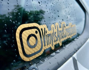 Może przedstawiać: Naklejka o fakturze złotego włókna węglowego umieszczona na szybie samochodu, przedstawiająca czarną ikonę logo Instagrama oraz czarny napis VinylsAndCreations. Naklejka znajduje się na szkle pokrytym drobnymi kroplami deszczu.