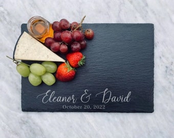 Puede incluir: Una tabla de servir de pizarra negra con una inscripción blanca que dice "Eleanor & David October 20, 2022". La tabla está decorada con uvas, fresas y una cuña de queso.