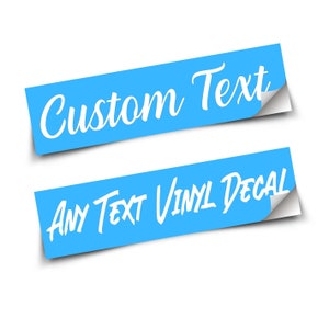 May include: Two rectangular blue vinyl decals with white text. The top decal features the words Custom Text in a cursive script. The bottom decal displays the words ANY TEXT VINYL DECAL in a bold, sans-serif font. Both stickers have a peeled corner.