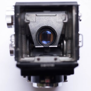 May include: Close-up of a vintage twin-lens reflex camera, showcasing the viewfinder and lens. The camera body is black and silver, with intricate mechanical details. The lens reflects a blue hue, and the camera is set against a white background.
