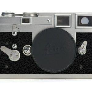 May include: A silver and black Leica rangefinder camera with a black lens cap. The camera has a textured black leather body and various dials and levers. The lens cap features the Leica logo.