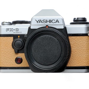 Può includere: Una fotocamera Yashica FX-D Quartz con parte superiore argentata e impugnatura color cuoio. L'obiettivo della fotocamera è nero e il marchio "Yashica" è stampato in argento. È visibile anche il modello "FX-D Quartz".