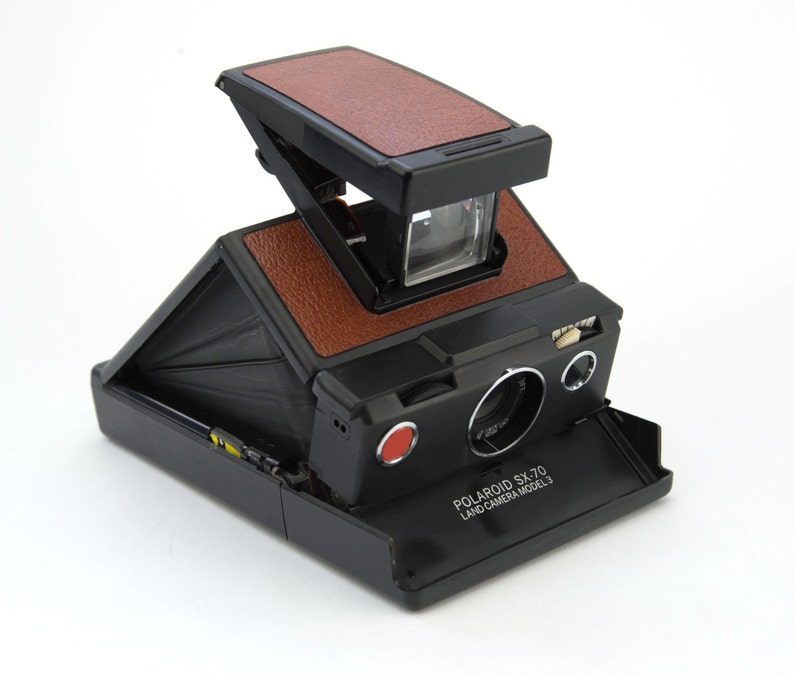 Puede incluir: Una c&aacute;mara Polaroid SX-70 Land Camera Model 3 vintage, negra con detalles en cuero marr&oacute;n. La c&aacute;mara tiene un dise&ntilde;o plegable, un bot&oacute;n rojo para disparar y una lente. El texto "POLAROID SX-70 LAND CAMERA MODEL 3" es visible.