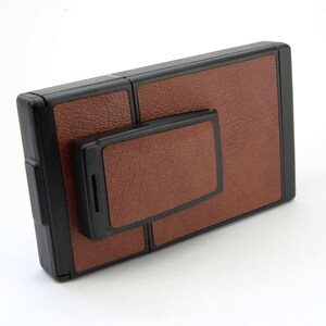 Puede incluir: Una c&aacute;mara Polaroid vintage con un acabado marr&oacute;n similar al cuero y bordes negros. La c&aacute;mara tiene una forma rectangular con un componente m&aacute;s peque&ntilde;o adjunto. El dise&ntilde;o es cl&aacute;sico y retro, enfocado en la fotograf&iacute;a instant&aacute;nea.