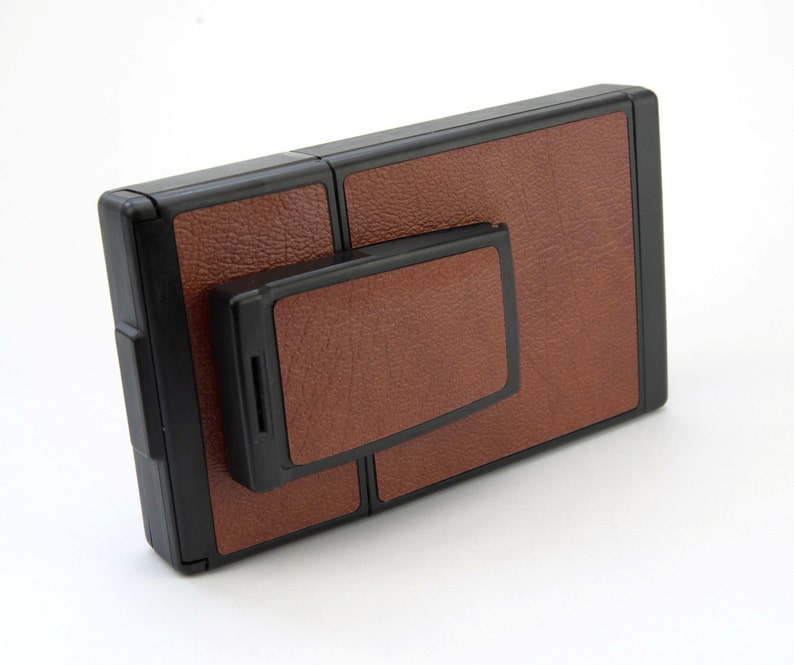 Puede incluir: Una c&aacute;mara Polaroid vintage con un acabado marr&oacute;n similar al cuero y bordes negros. La c&aacute;mara tiene una forma rectangular con un componente m&aacute;s peque&ntilde;o adjunto. El dise&ntilde;o es cl&aacute;sico y retro, enfocado en la fotograf&iacute;a instant&aacute;nea.