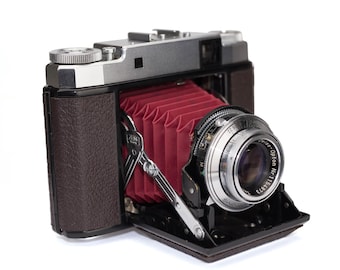 Puede incluir: Una cámara plegable vintage de plata y negro con fuelle rojo y una lente plateada con la inscripción "Optron Nr 1 1:3.5 74.5 mm 154873".