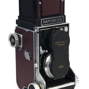 May include: A vintage Mamiyaflex camera with a maroon leather exterior and black accents. The camera features a silver lens and the text "MAMIYAFLEX" and "MAMIYAFLEX-C Professional". The camera has a classic, boxy design.