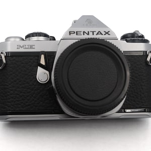Puede incluir: Una cámara de película Pentax ME plateada con una tapa de lente negra. La cámara tiene una cubierta de imitación de cuero negra y una anilla de correa plateada en el lado izquierdo.
