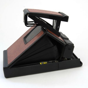 Puede incluir: Una c&aacute;mara Polaroid vintage con cuerpo negro y paneles texturizados marrones. La c&aacute;mara est&aacute; abierta, revelando el fuelle y los componentes internos. El dise&ntilde;o incluye un visor y una montura para flash.