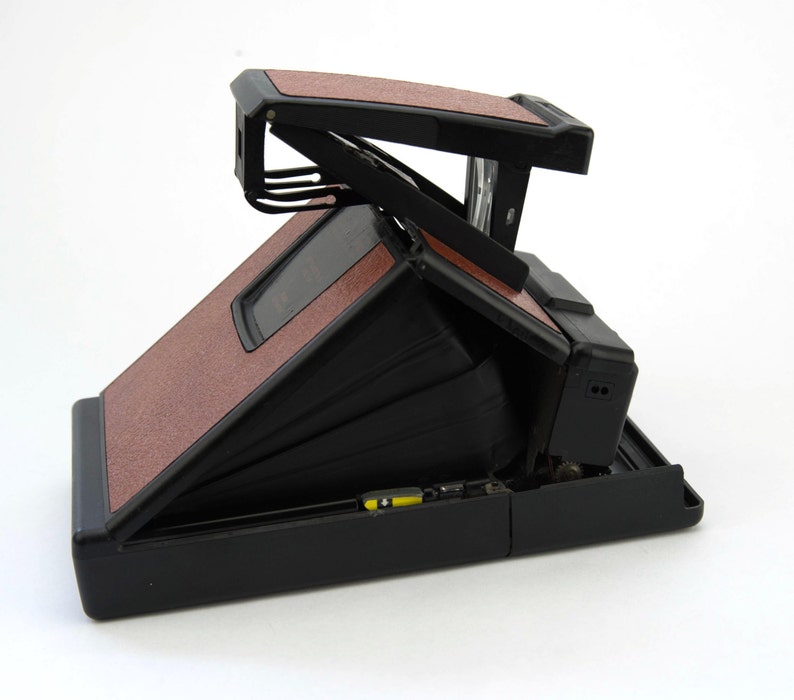 Puede incluir: Una c&aacute;mara Polaroid vintage con cuerpo negro y paneles texturizados marrones. La c&aacute;mara est&aacute; abierta, revelando el fuelle y los componentes internos. El dise&ntilde;o incluye un visor y una montura para flash.
