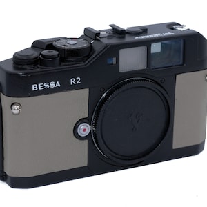 Può includere: Una fotocamera a telemetro BESSA R2 nera e grigia con un tappo dell'obiettivo nero. La fotocamera ha una vite argentata sul lato e un punto rosso sul davanti.