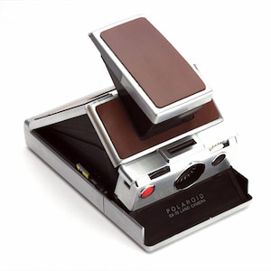 Peut inclure: Un appareil photo Polaroid SX-70 Land Camera argenté et brun avec le cache de l'objectif ouvert. L'appareil photo a un bouton rouge et une étiquette noire qui indique "POLAROID SX-70 LAND CAMERA".
