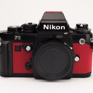 Puede incluir: Una cámara de película Nikon F3 negra y roja con una tapa de lente negra. La cámara tiene una inscripción plateada que dice "F3" y "HP".