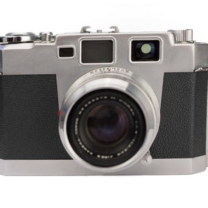 Puede incluir: Una cámara de película de 35 mm de color plateado y negro con una lente y un visor. La cámara tiene una cubierta de cuero en el cuerpo.