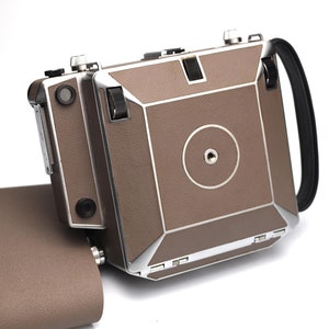 Peut inclure: Un appareil photo vintage brun et argent avec une sangle en cuir. L'appareil photo a un cache d'objectif rond et un viseur rectangulaire.