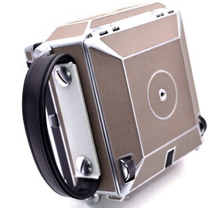 Puede incluir: Una cámara de formato medio vintage con un cuerpo marrón y plateado y un asa de cuero negro. La cámara tiene forma cuadrada con paneles geométricos y un detalle de lente circular. La cámara está en ángulo, mostrando su lado y la parte superior.