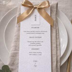 May include: A formal table setting featuring a menu card with a gold ribbon. The menu card lists a starter, main course, and dessert. The place setting includes white plates, a linen napkin, and gold cutlery. The menu is for Darcy.