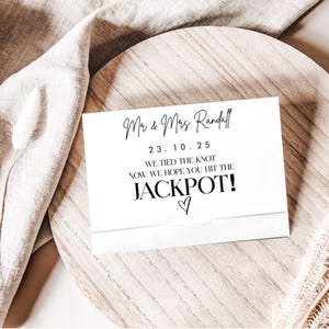 Könnte beinhalten: Eine weiße Karte mit schwarzem Text, der "Mr & Mrs Randall", ein Datum und "WE TIED THE KNOT NOW WE HOPE YOU HIT THE JACKPOT!" mit einem Herzsymbol enthält. Die Karte liegt auf einer Holzoberfläche.