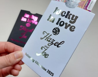 Puede incluir: Sobre de cartulina blanca con un diseño de lámina plateada que dice "lucky in love Hazel and Joe" y "Saturday 11th June 2025". El sobre tiene una ranura para una tarjeta o dinero y un boleto de lotería es visible en el interior.