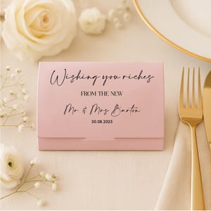 Könnte beinhalten: Eine hellrosa Hochzeitsgeschenkbox mit dem Text "Wishing you riches" und "Mr & Mrs Barton 30.08.2025". Die Box befindet sich auf einer Tischdekoration mit goldenem Besteck und weißen Blumen.