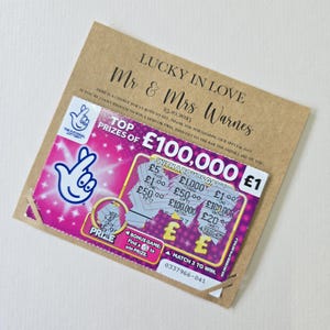 Puede incluir: Una tarjeta marrón con el texto "LUCKY IN LOVE" y "Mr & Mrs Warnes" impreso. Se adjunta un billete de lotería rosa para rascar, con un premio mayor de £100.000. La tarjeta incluye la fecha 25.10.2025 y un mensaje.