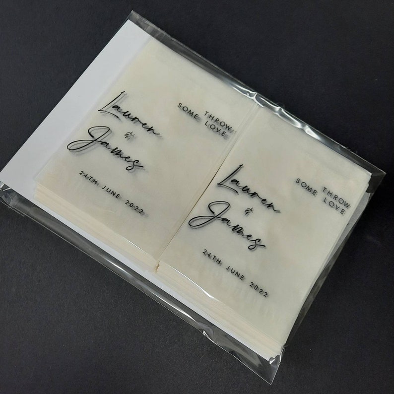 May include: A stack of white tissue paper favors with black text that reads "Lauren & James" and "Some Throw Love" with the date "24th June 2022".