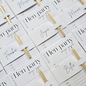 K&ouml;nnte beinhalten: Wei&szlig;e Karten mit dem Text "Hen party emoji game" und Namen in blauer Schrift. Jede Karte hat einen Holzstift. Die Karten sind auf einer hellen Oberfl&auml;che angeordnet, f&uuml;r ein Partyspiel.