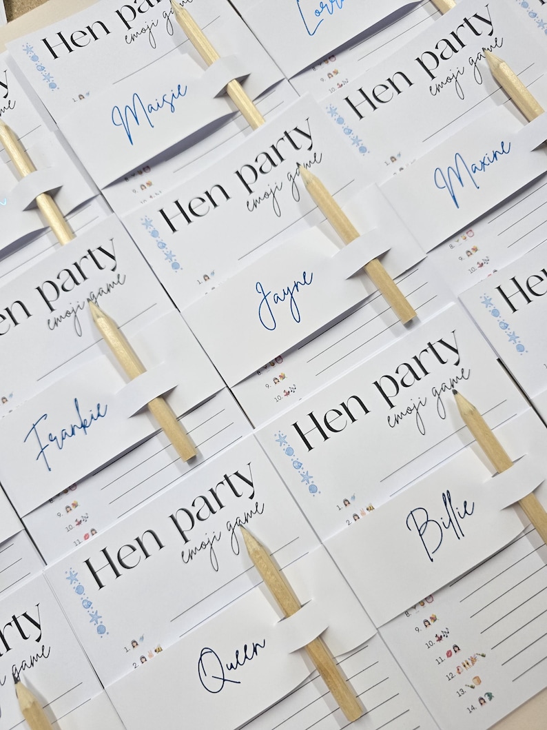 K&ouml;nnte beinhalten: Wei&szlig;e Karten mit dem Text "Hen party emoji game" und Namen in blauer Schrift. Jede Karte hat einen Holzstift. Die Karten sind auf einer hellen Oberfl&auml;che angeordnet, f&uuml;r ein Partyspiel.