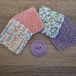 Peut inclure: Quatre dessous de verre faits à la main au crochet, dans les tons de rose, blanc, bleu et violet. Un bouton lavande avec le texte "handmade by hannah" est au centre. Les dessous de verre sont en fil doux et texturé.