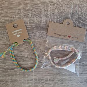 Può includere: Due braccialetti dell'amicizia fatti a mano. Uno ha un motivo a spina di pesce in blu, arancione e verde. L'altro è color pastello e confezionato in un sacchetto trasparente. Entrambi hanno etichette con la scritta "Handmade with love."