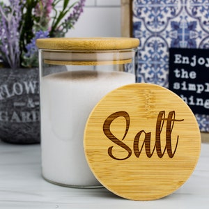 May include: A glass jar with a bamboo lid and a bamboo lid with the word "Salt" engraved on it. The jar is filled with white salt.