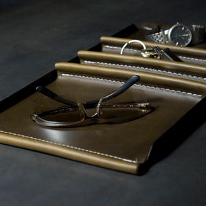 May include: A brown leather tray with four compartments, each with a stitched edge. The tray is empty except for a pair of sunglasses in the largest compartment.